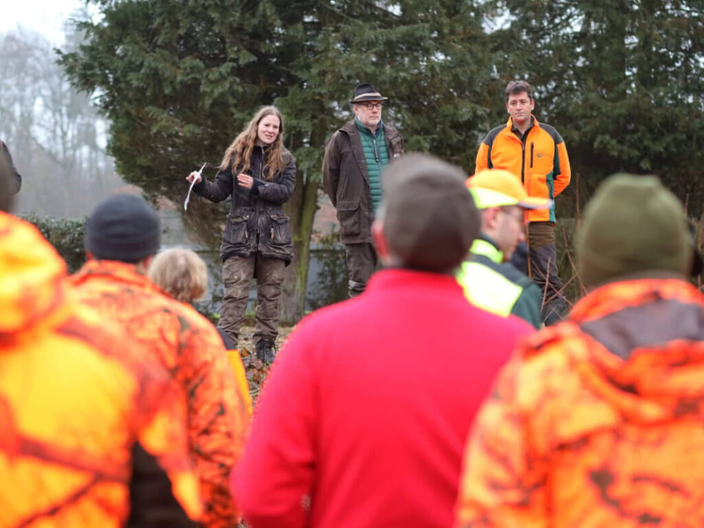 Eine größere Gruppe Menschen im Wald. Davon sieht man etwa sechs Personen in roter und gelber Warnkleidung von hinten. Sie schauen auf eine Gruppe aus einer Frau und zwei Männern in graugrüner Kleidung. Die Frau erklärt der größeren Gruppe etwas.