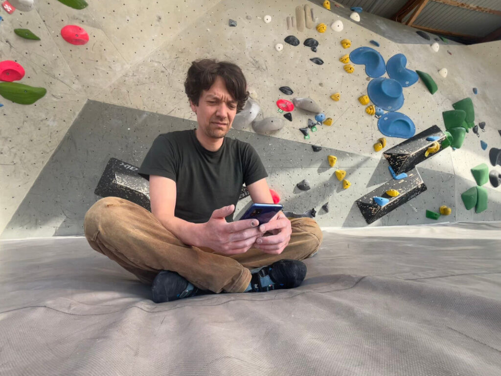 Eine Kletterhalle. Im Hintergrund sieht man eine Kletterwand, im Vordergrund sitzt auf den Matten der Halle ein Mann. Er hat ein Smartphone in den Händen und betrachtet dessen Display.