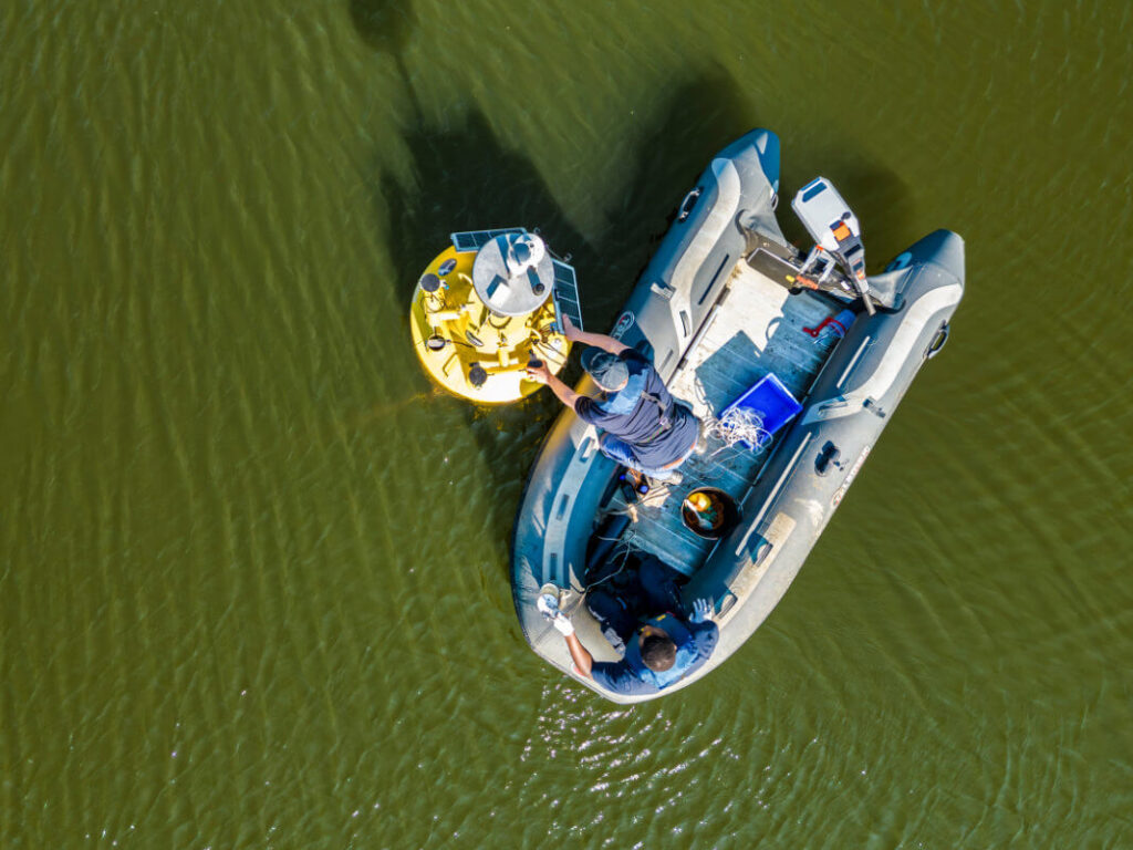Drohnenansicht einer im Sonnenlicht glänzenden Seeoberfläche. Auf dem See schwimmen eine Boje, die mit zahlreichen technischen Gerätschaften ausgestattet ist und direkt daneben ein Schlauchboot mit Heckmotor. Im Schlauchboot befinden sich zwei Personen. Eine der Personen wartet die Geräte auf der Boje.